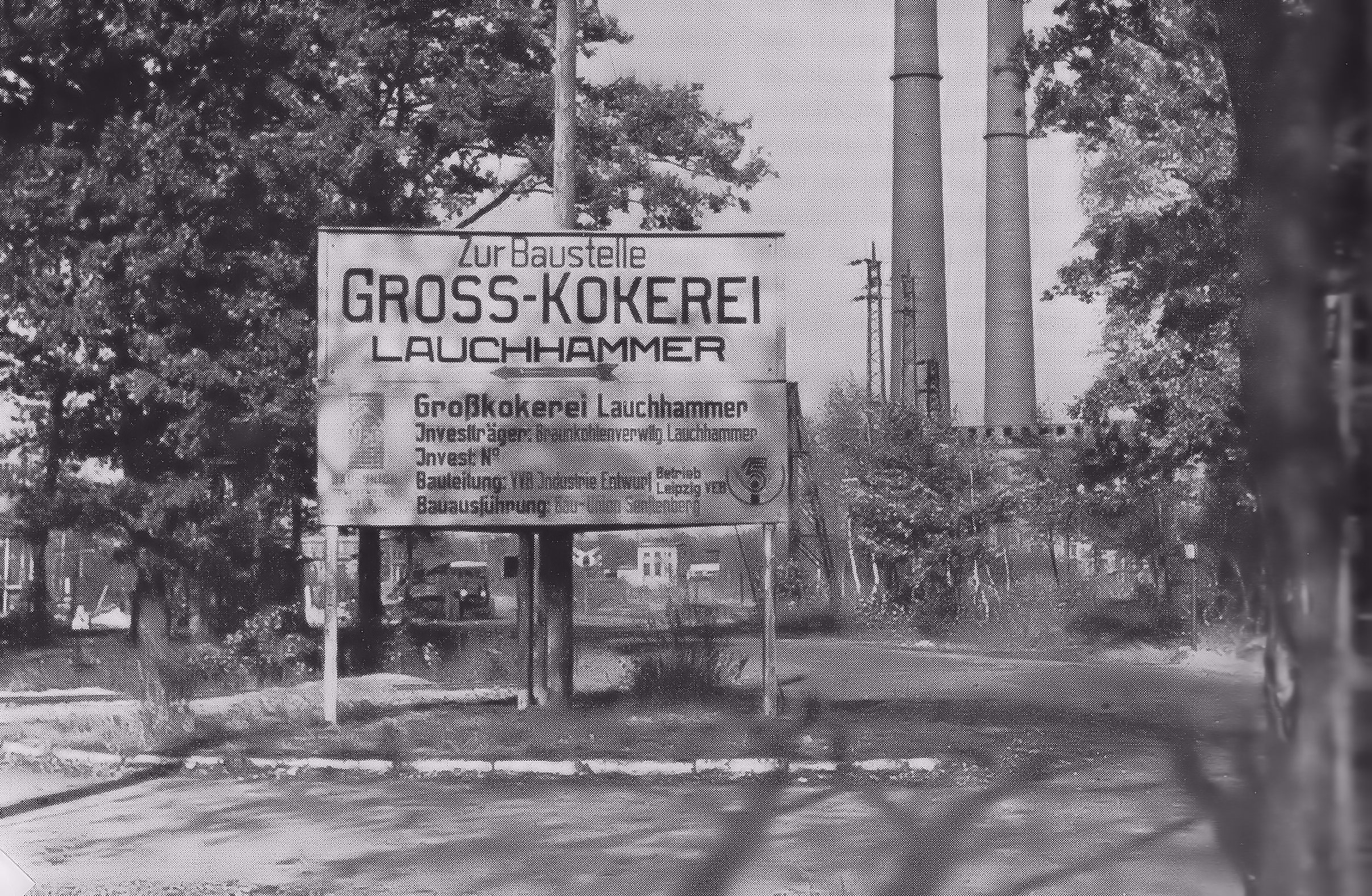 Blick auf den Eingang zur Baustelle der Gross-Kokerei in Lauchhamemer. Auf einem Schildt steht: "Zur Baustelle Gross-Kokerei Lauchhammer". Im Hintergrund sind zwei Schlote und Bäume zu sehen. Bildquelle: Heimatverein Lauchhammer.