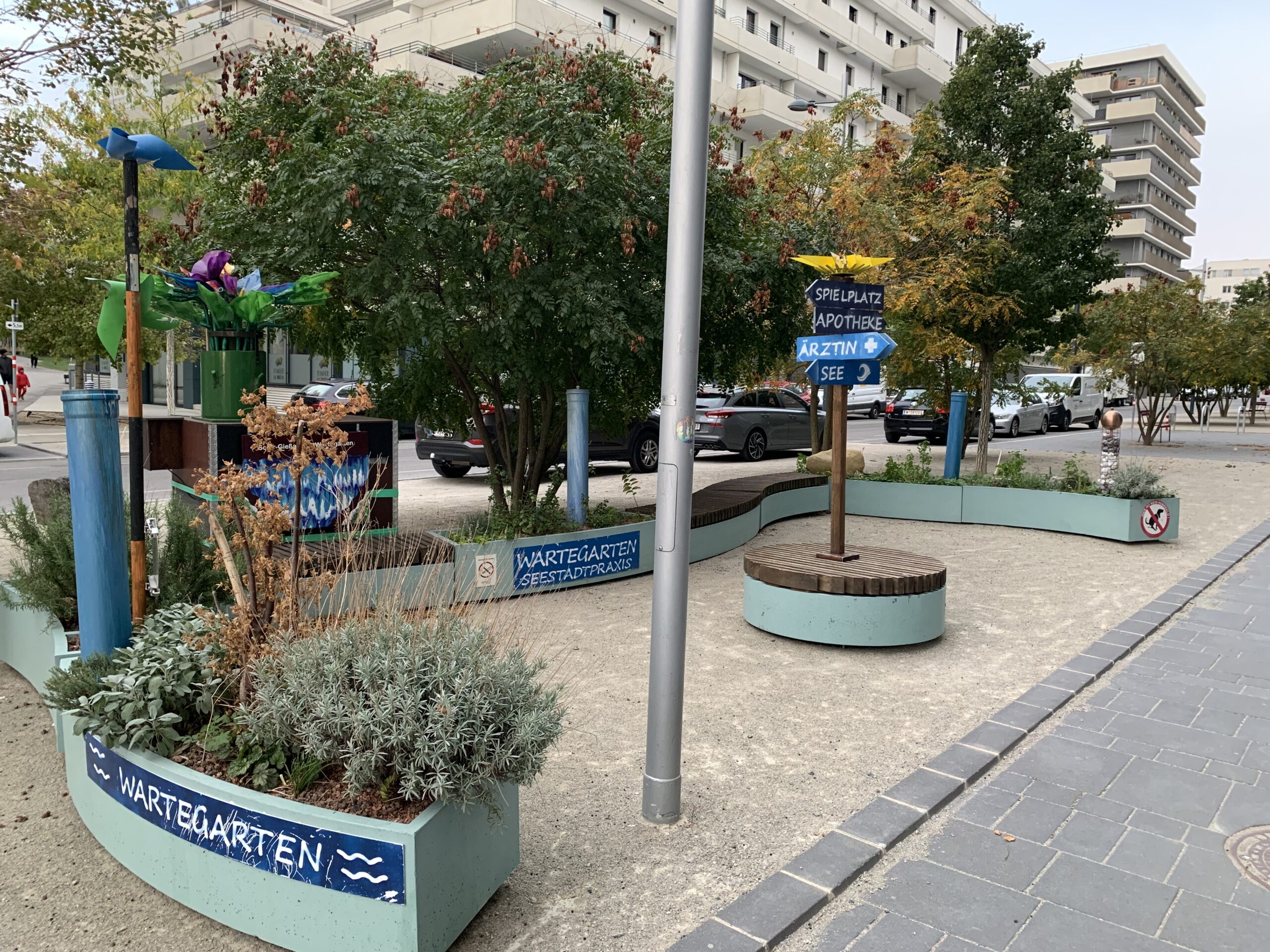 Ein Platz mit Sitzbändken, bepflanzten Beeten und Bäumen. In der Mitte steht ein Wegweiser, in welche Richtung es zum Arzt, zum See und zur Apotheke geht. Im Hintergrund Häuser.