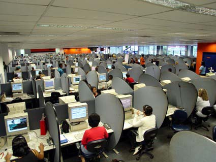 Ein Call Centre in Südasien: Menschen sitzen nebeneinander gereihnt an einem Tisch. Die Arbeitsbereiche der einzelnen Personen sind durch Raumtrenner geteilt. In jedem Arbeitsbereich steht ein Computer.