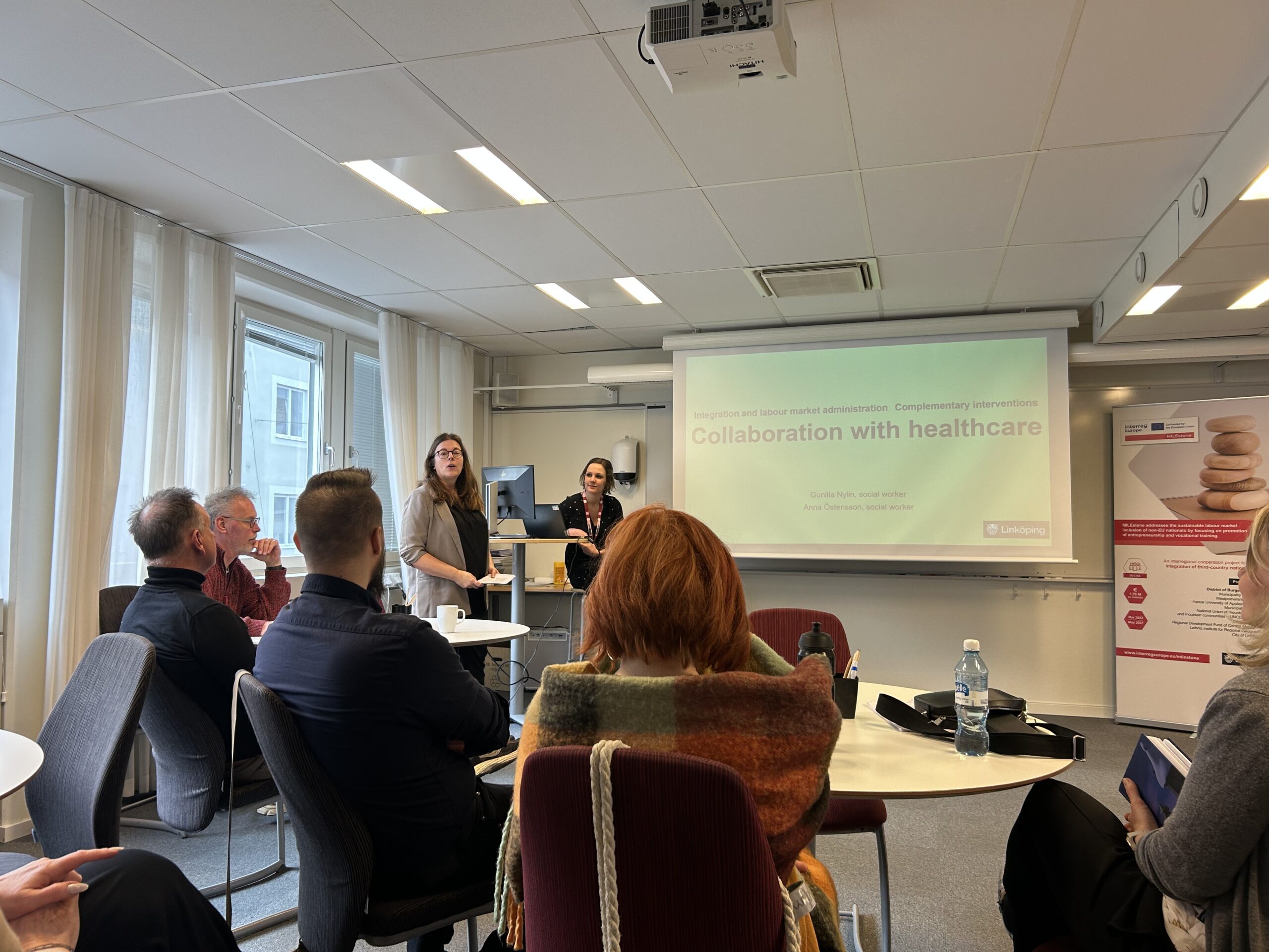 Eine Gruppe Menschen sitzt in einem Seminarraum und hört einem Vortrag von zwei schwedischen Frauen zu, die über Möglichkeiten der Gesundheitsfürsorge für Migrant:innen sprechen.