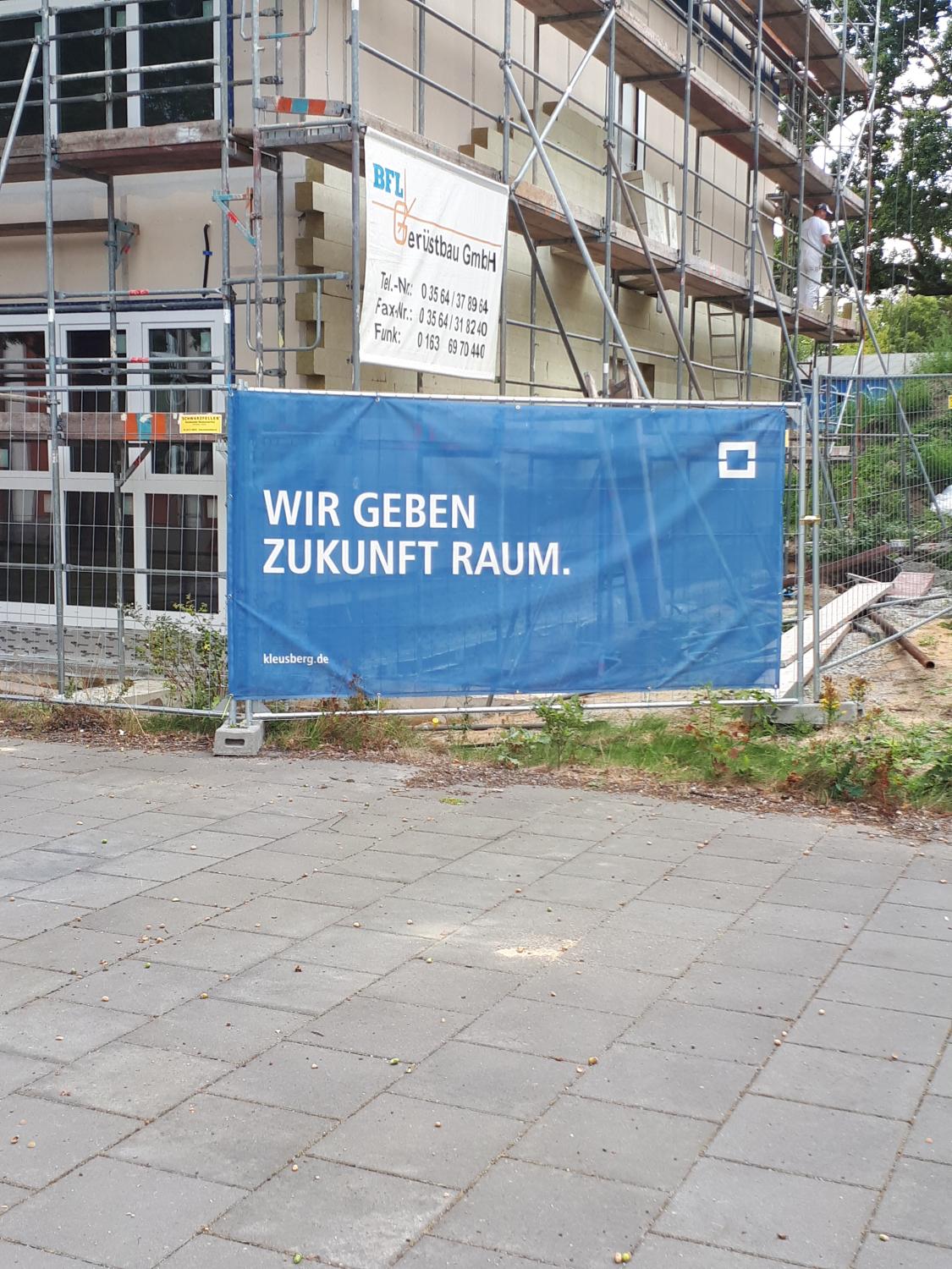 Ein Haus mit einem Baugerüst. Davor sperr ein Absperrzaun den Zugang ab. Am Zaun hängt ein Banner mit der Aufschrift: "Wir geben Zukunft Raum."