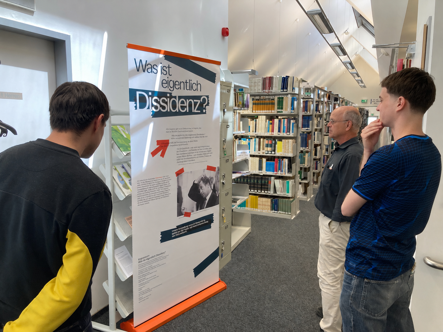 Drei Personen schauen sich ein Poster der Ausstellung "Was ist eigentlich Dissidenz?" in der Bibliothek des GWZO in Leipzig an.