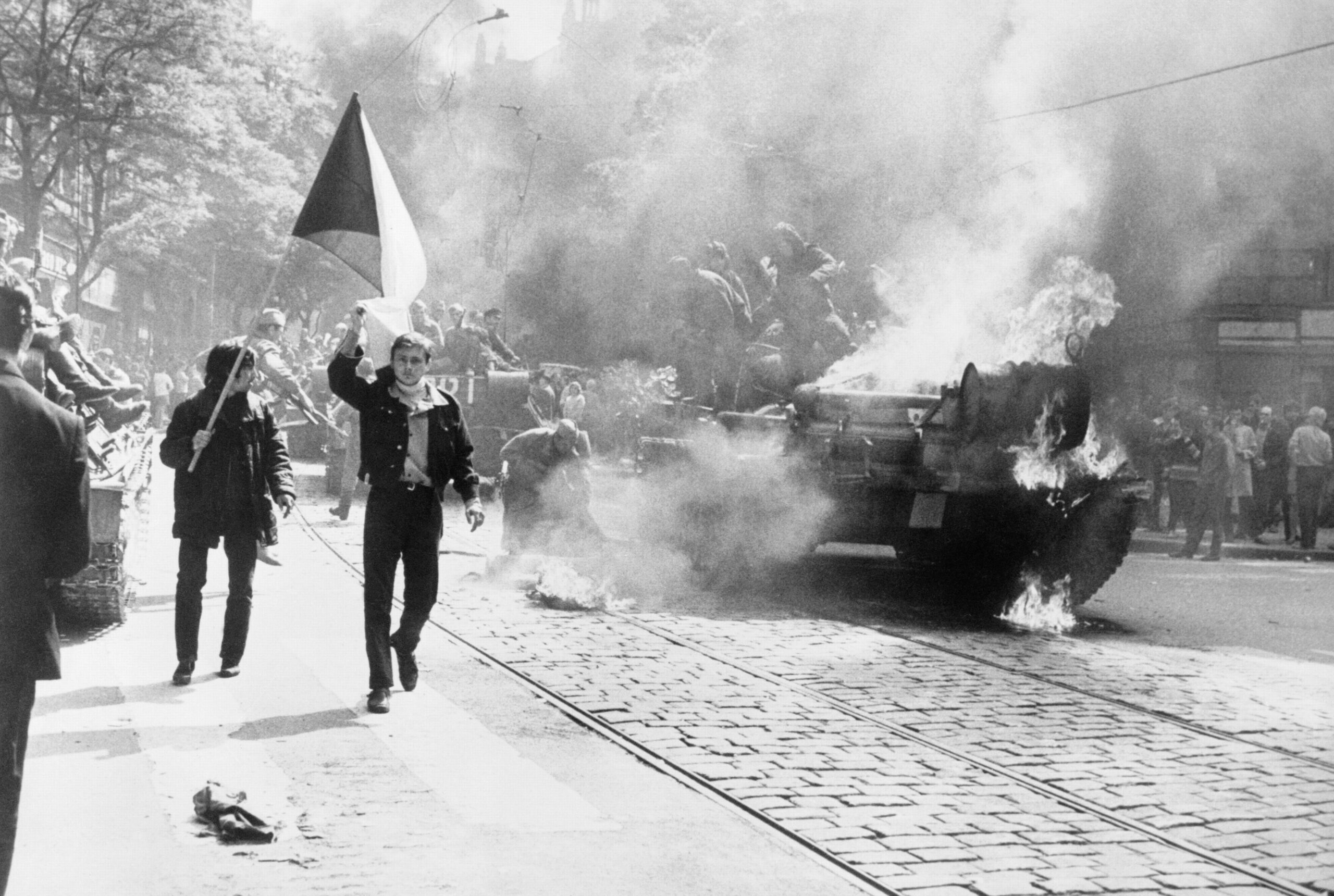 Aufnahme w&auml;hrend der Besatzung Prags durch milit&auml;rische Truppen des Warschauer Pakts. Auf einer Stra&szlig;e steht ein brennender Panzer. Daneben laufen M&auml;nner mit Tschechischer Flagge in der Hand.