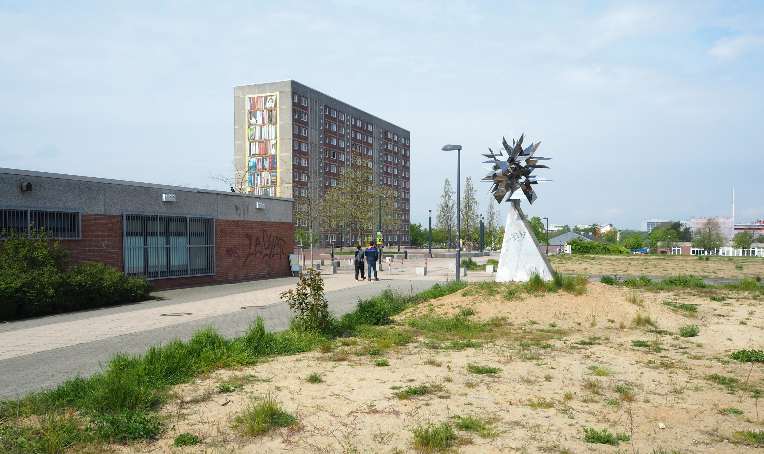 Der Berliner Platz in Schwerin: Eine weite, karge Fläche im Vordergrund, dahinter ein Flachbau und ein Plattenbau. Am Rand des Platzes steht eine Kunstinstallation.