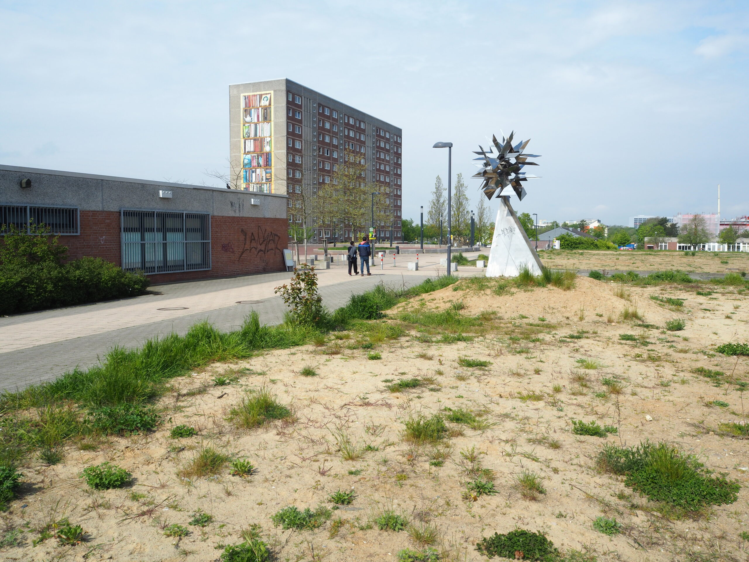 Der Berliner Platz in Schwerin: Eine weite, karge Fl&auml;che im Vordergrund, dahinter ein Flachbau und ein Plattenbau. Am Rand des Platzes steht eine Kunstinstallation.