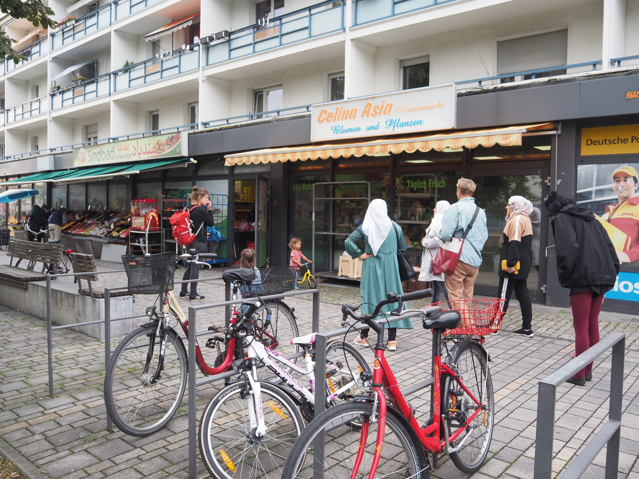 Menschen laufen an einem Plattenbau vorbei, in dessen Erdgeschoss sich Gesch&auml;fte des t&auml;glichen Bedarfs befinden. Davor stehen Fahrradst&auml;nder mit angeschlossenen R&auml;dern.