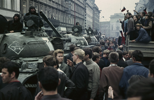 Aufnahme vom Prager Fr&uuml;hling 1968: Mehrere Panzer stehen hintereinander gereiht in einer Stra&szlig;e voller menschen. Rechts im Bild steht ein Fahrzeug mit Anh&auml;nger, auf dem Menschen die teschische Flagge wehen.