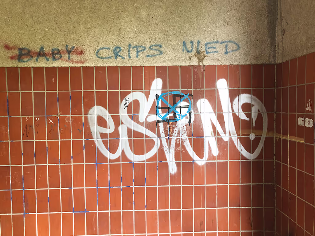 Graffiti auf einer Wand in einem abgenutzten &ouml;ffentlichen Geb&auml;ude. Auf einem nicht n&auml;her bekannten Tag ist ein Hakenkreuz in Schwarz gesprayt, das mit blauer Farbe durchgestrichen wurde.
