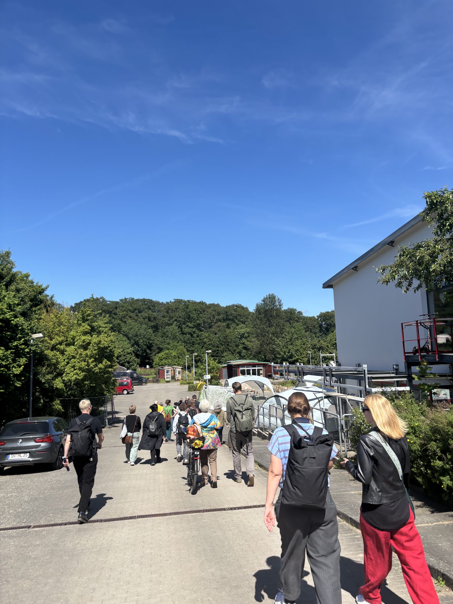 Die Teilnehmer:innen des Stadtspaziergangs laufen &uuml;ber eine Stra&szlig;e. Rechts davon steht ein gr&ouml;&szlig;erer Neubau, links davon B&auml;ume.